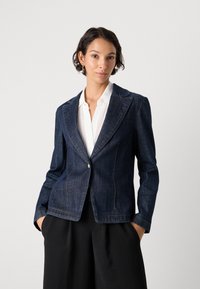 Mørkeblå denimblazer med en skreddersydd passform, to frontlommer og kontraststing. Brukt over en hvit skjorte med krage.