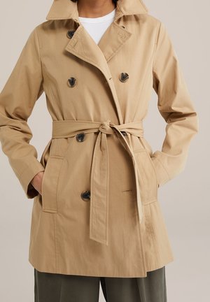 Vrouw die een beige double-breasted trenchcoat draagt met donkere knopen, een gestrikte riem en handen in de voorzakken over een wit overhemd en donkere broek.