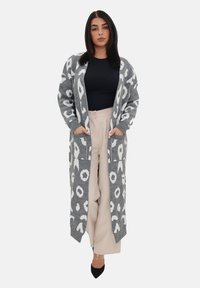 Cardigan lungo grigio lavorato a maglia con macchie leopardate bianche, dotato di un colletto sciallato, tasche laterali e polsini a coste. Indossato sopra un top nero.