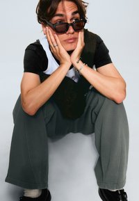 Tricou supradimensionat negru și gri, stratificat cu o cămașă albă. Pantaloni gri largi, pantofi negri și ochelari de soare dreptunghiulari maro. Fundal deschis.