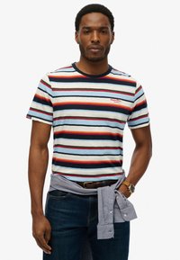 Stribet t-shirt i blå, rød og creme med en marineblå krave, prydet med et lille rødt logo på brystet; iført med mørke jeans og en bundet skjorte.