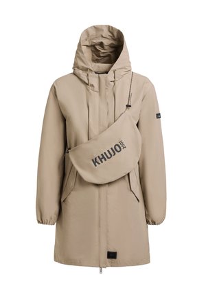 Frau steht, trägt eine beige Kapuzenjacke über passenden Leggings und Turnschuhen, schaut nach vorne vor einem schlichten grauen Hintergrund.