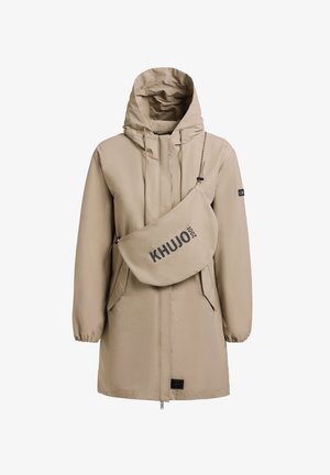 Vrouw staand, gekleed in een beige gewatteerde jas met capuchon, gecombineerd met bijpassende legging en sneakers, kijkt recht vooruit tegen een effen grijze achtergrond.