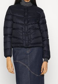 Žena v černé péřové bundě s předními kapsami a tmavých džínových sukních s zakřiveným prošíváním, stojící na jednolitém pozadí.