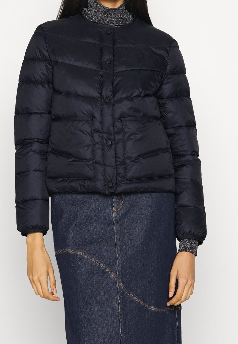 Žena v černé péřové bundě s předními kapsami a tmavých džínových sukních s zakřiveným prošíváním, stojící na jednolitém pozadí.