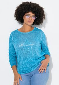 Maglia a manica lunga color turchese con scollatura rotonda, caratterizzata da un motivo a stella in paillettes sulla parte anteriore e tessuto testurizzato. Abbinata a jeans azzurri chiari.