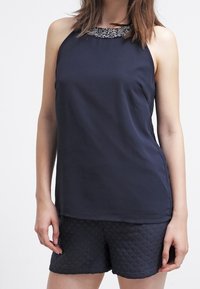 Marinblå ärmlös topp med pärlmandel, i kombination med mörka vadderade shorts. Slät tyg, avslappnad passform och minimalistisk design.