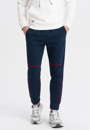 Mørkeblå sweatpants med røde syninger, tapered fit, ribbet tekstur og elastiske manchetter, parret med lyse grå og hvide sneakers.