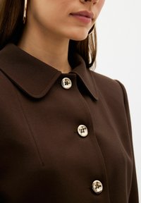 Brun knäppt jacka med en strukturerad krage, metallknappar och slät tygtextur. Närbilden framhäver detaljrikedom i hårdvaran.