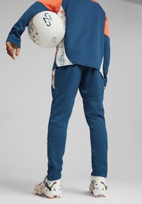 Ropa deportiva en azul con detalles en naranja. Parte superior y pantalones texturizados, sosteniendo un balón de fútbol con un diseño estampado. El diseño incluye elementos gráficos en el lado.