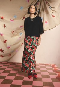 Pulover negru pufos, fustă maxi cu model floral cu trandafiri roșii pe fond negru și pantofi roșii cu vârf ascuțit. Fundalul prezintă draperii cu model de stele.