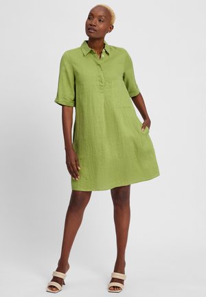 Green linen shirt dress with a collar, short rolled sleeves, a chest pocket, and a relaxed fit. Worn with light-colored sandals.
