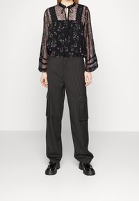 Femme portant un chemisier transparent noir à fleurs avec des détails en dentelle, un pantalon cargo à fines rayures, et des chaussures à plateforme en cuir verni noir, debout sur un sol blanc.