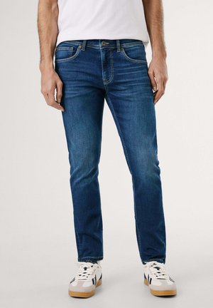 Homme portant un jean slim bleu foncé et des baskets blanches avec des détails noirs, debout sur un fond clair uni.