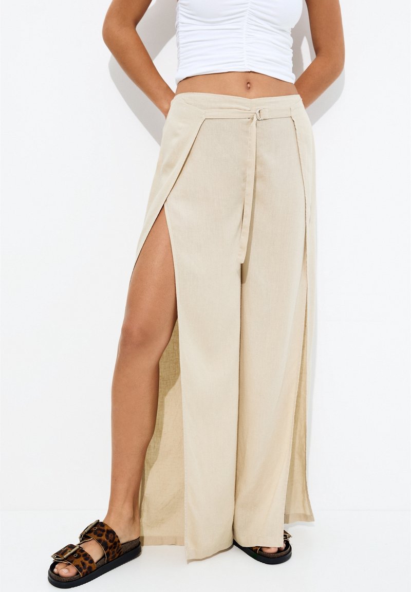 Femme portant un pantalon large beige avec de hautes fentes latérales, un débardeur blanc et des sandales à boucle imprimé léopard.