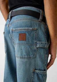 Jasnoniebieskie dżinsy z denimu z naszywką ze skóry oznaczoną napisem "GUESS JEANS 1981", wyposażone w wiele kieszeni i detale z kontrastującym przeszyciem.
