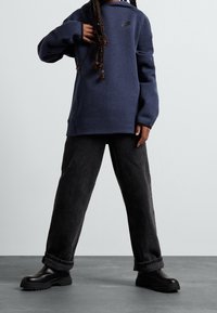 Marinblå oversized sweatshirt med en liten svart logo, kombinerad med löst sittande svarta denimbyxor och kraftiga svarta stövlar.