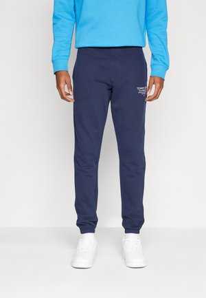 Hombre con joggers azul marino, zapatillas blancas y una sudadera azul brillante, de pie frente a un fondo claro y liso.