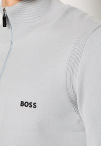 Sweater zippé gris clair fabriqué en tissu tricoté, avec un col haut et un subtil motif côtelé. Logo "BOSS" noir sur la poitrine.