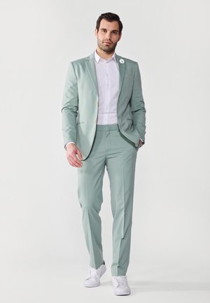 Light green suit with tailored fit, single-breasted jacket, white dress shirt, and white sneakers. Features a decorative lapel pin.