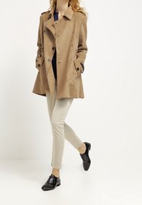 Beżowy płaszcz trench z zamszu o podwójnym zapięciu, srebrnymi guzikami i kieszeniami bocznymi, zestawiony z beżowymi spodniami i czarnymi butami.