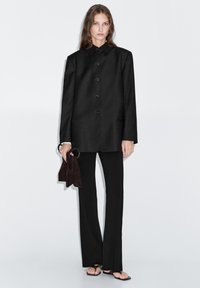 Black tailored coat with a high collar, button closure, and two front pockets, paired with black flared pants and a dark suede handbag.