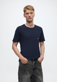 T-shirt di cotone blu navy con maniche corte, scollo rotondo e taschino sul petto con piccolo logo giallo, abbinata a jeans neri sbiaditi.