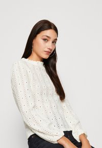 Balta teksturēta blūze ar viļņotu apkakli, garām puffētām piedurknēm un mazām izgriezumu detaļām, apvienota ar tumšām džinsa biksēm.