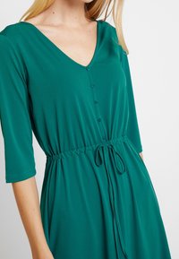 Robe teal avec un décolleté en V, manches trois-quarts, bouton sur le devant et taille à cordon. Tissu lisse et léger avec une texture douce.
