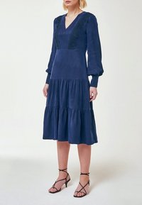 Mörkblå klänning i mjuk tyg, med V-ringning, puffärmar och en skiktad kjol. Utsmyckad med spetsdetaljer längs livstycket.