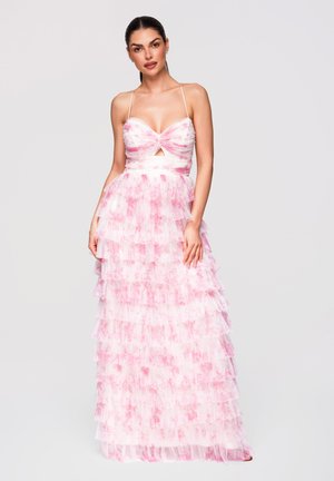 Femme portant une robe sans manches rose et blanche à volants étagés avec bretelles fines et une petite découpe à l'avant, debout devant un fond uni.