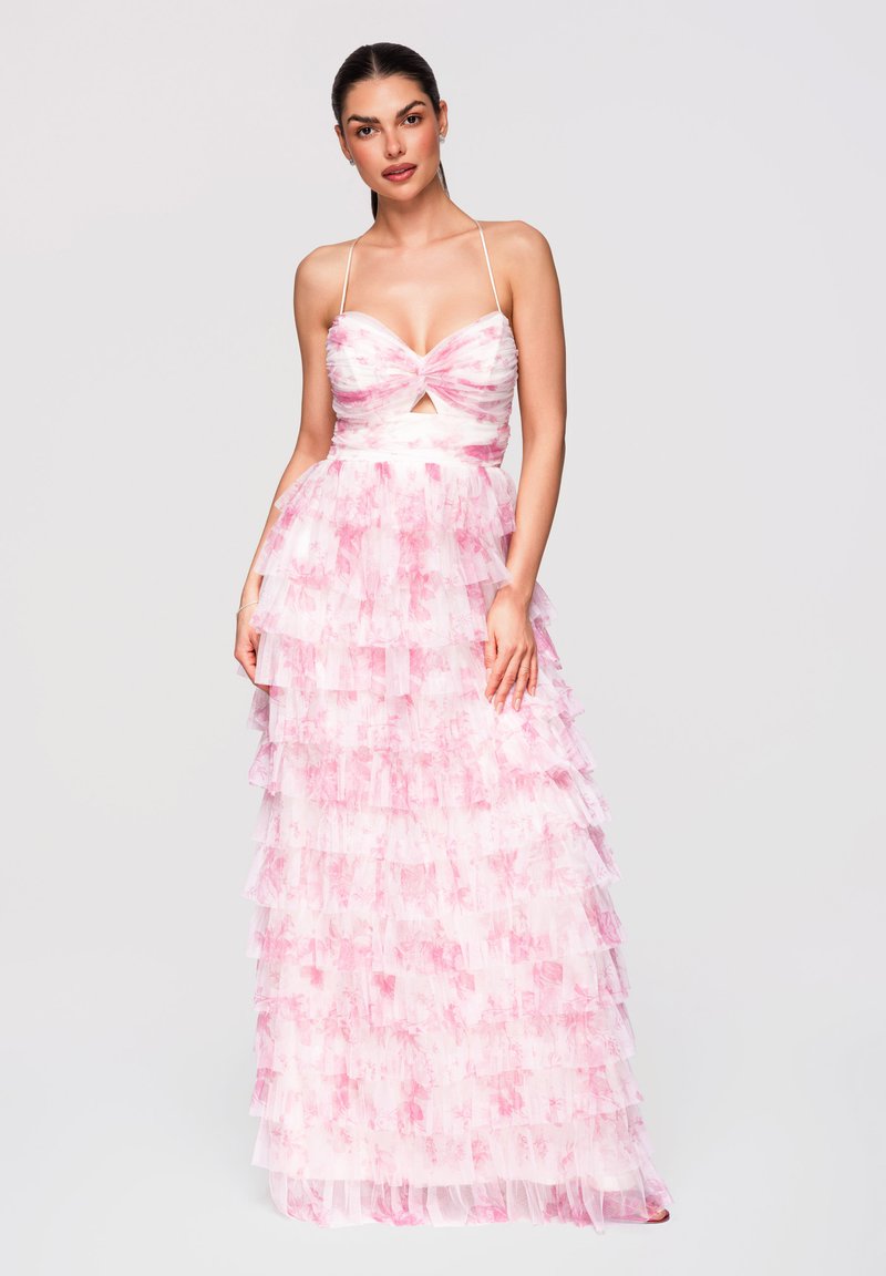 Femme portant une robe sans manches rose et blanche à volants étagés avec bretelles fines et une petite découpe à l'avant, debout devant un fond uni.