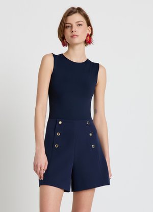 Donna che indossa una tuta blu navy senza maniche con bottoni dorati sul davanti e orecchini con piume rosse, in piedi davanti a uno sfondo chiaro e uniforme.