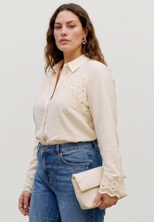 Une femme porte un chemisier crème à bordure en dentelle, un jean bleu, des créoles dorées et une pochette assortie sur un fond uni.