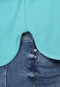 Türkises kurzärmliges Oberteil mit abgerundetem Saum, das über ausgewaschenen blauen Denim-Jeans getragen wird, die einen sichtbaren Metallknopf und eine distresste Textur aufweisen.