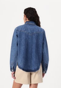 Jachetă din denim de culoare albastru mediu, cu o umeră ascuțită, mâneci lungi, manșete cu nasturi și un tiv curbat. Textură aspră cu cusături vizibile.
