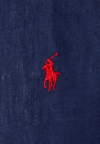 Námořnická tkanina s texturovanou vazbou, která má výrazné červené vyšívané logo poníka, zobrazující jezdeckého muže s hůlkou.