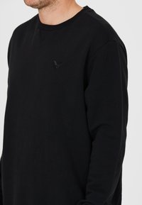 Svart långärmad sweatshirt med rundad halsringning och textilstruktur. Har en liten svart logotyp på vänster bröstområde.