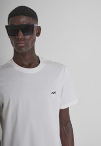 Camiseta blanca de algodón con cuello redondo y mangas cortas, que presenta una pequeña etiqueta de logo negra en el pecho. Gafas de sol negras de gran tamaño.