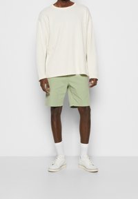Maglietta a maniche lunghe a coste crema, pantaloni corti verde chiaro, calze bianche, sneakers bianche. Design semplice, vestibilità rilassata, colori moderni.