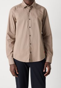 Hombre vestido con una camisa de botones beige de manga larga y pantalones negros, de pie contra un fondo claro y liso.