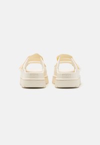 Beige sandalen met een chunky zool, open achterkant en verstelbare banden. UGG-logo zichtbaar op de hak. Gladde textuur en minimalistisch ontwerp.