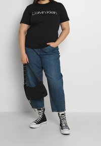 Camiseta negra con el logo de "Calvin Klein", combinada con pantalones vaqueros de tiro alto azules y zapatillas altas negras, sosteniendo un bolso negro de hombro.