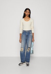 Bluză lungă cu mânecă din tricot crem, blugi ripitați de un albastru deschis, adidași alb-negru și o geantă crossbody de un albastru deschis. Design simplu, stil casual.