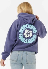 Lilla hættetrøje med et stort hvidt og lyseblåt blomsterlogo på ryggen, parret med lyseblå denim jeans.