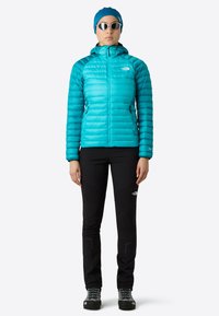 Turkos isolerad jacka med huva, svarta åtsittande byxor och klätterskor. Materialet är glänsande, med en puffig design och två framlommer.