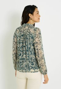 Blouse transparente à manches longues avec un fond bleu-vert présentant des motifs floraux beiges et des accents. Col boutonné et détail froncé au dos.