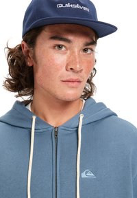 Sweat à capuche zippé bleu avec des cordons blancs et un petit logo blanc sur la poitrine gauche, porté avec une casquette marine arborant une marque blanche.