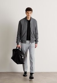 Giacca bomber grigia con zip, camicia nera, pantaloni grigio chiaro, scarpe nere e borsa in pelle nera. Texture lisce, design casual.