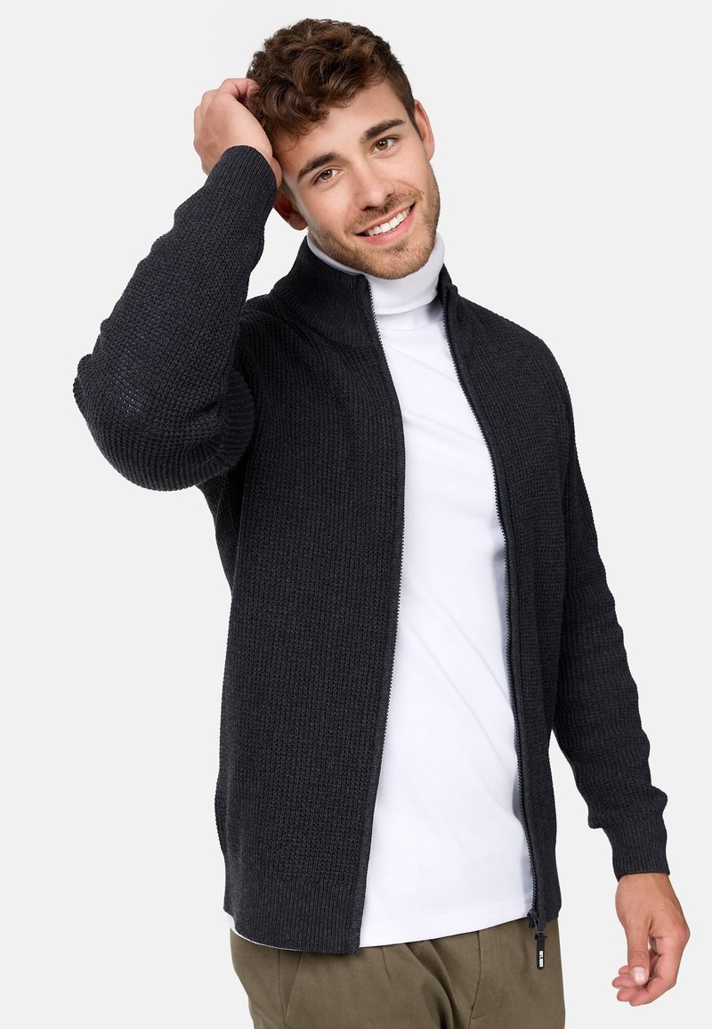 Mørkegrå strikket zip-up genser med høy krage, strukturert stoff og ribbestrikkede ermer, båret over en hvit turtleneck.